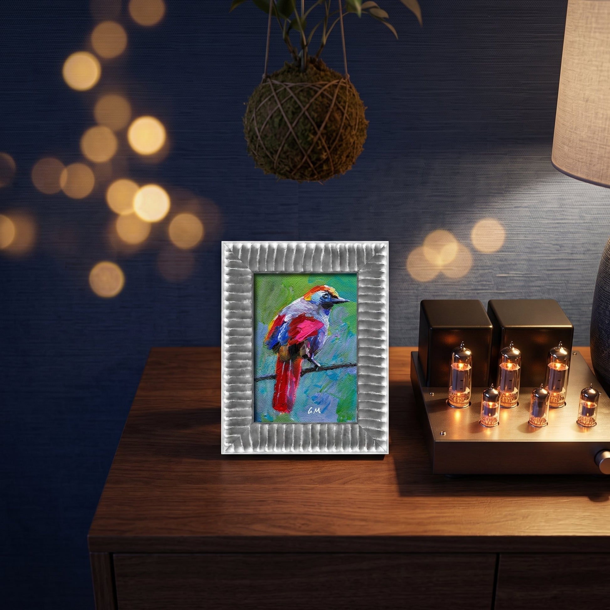 A colorful expressionist bird painting perched on a wooden console beside glowing bulbs and a hanging plant, framed in silver.
