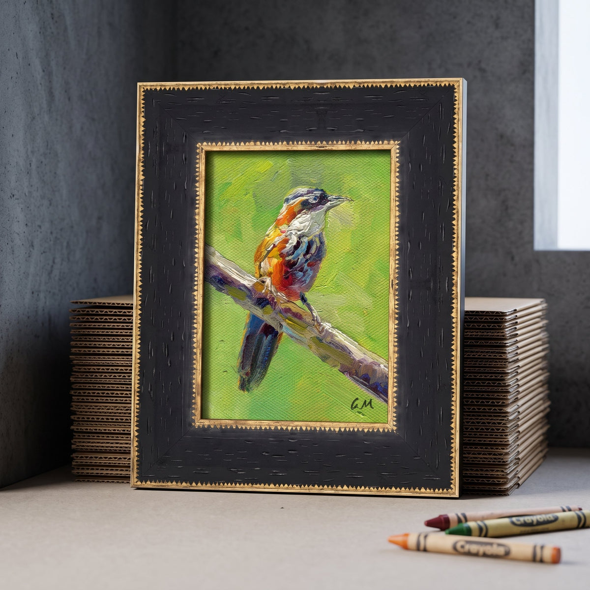 A vividly painted bird perched on a branch, set in a textured black frame with gold trim, rests on a worktable beside crayons and cardboard stacks.