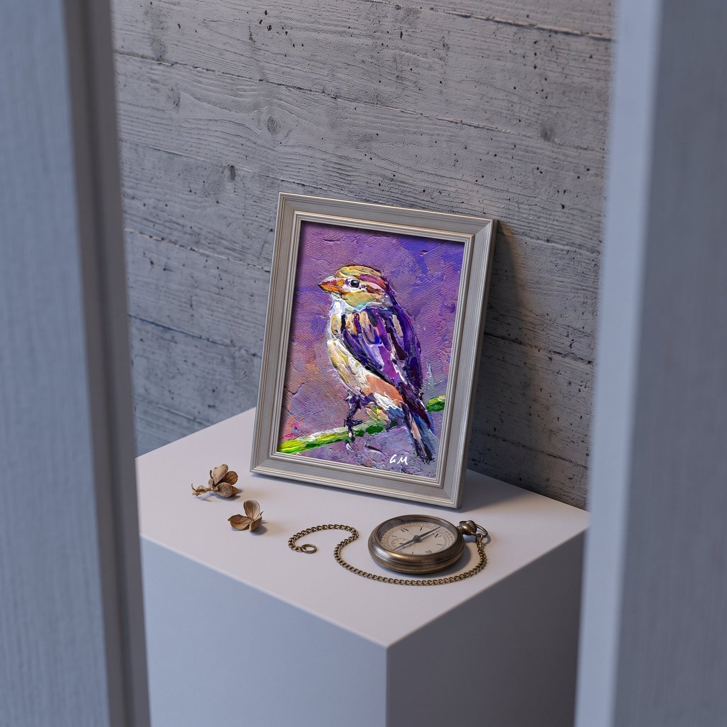 A colorful bird painting in purple, yellow, and white rests on a white pedestal beside a vintage pocket watch, framed in silver against a weathered wood wall.