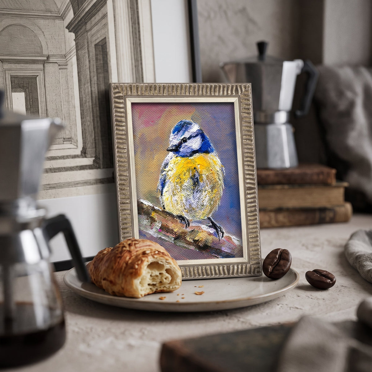 A vivid oil painting of a blue and yellow bird perched on a branch, framed in silver, sits on a kitchen counter beside a croissant and coffee beans.