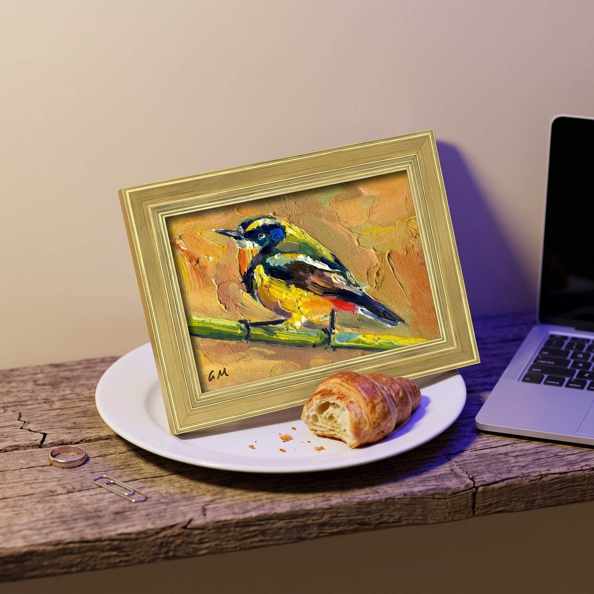 A golden-framed oil painting of a colorful bird perched on a branch sits on a white plate beside a croissant on a weathered wooden desk.
