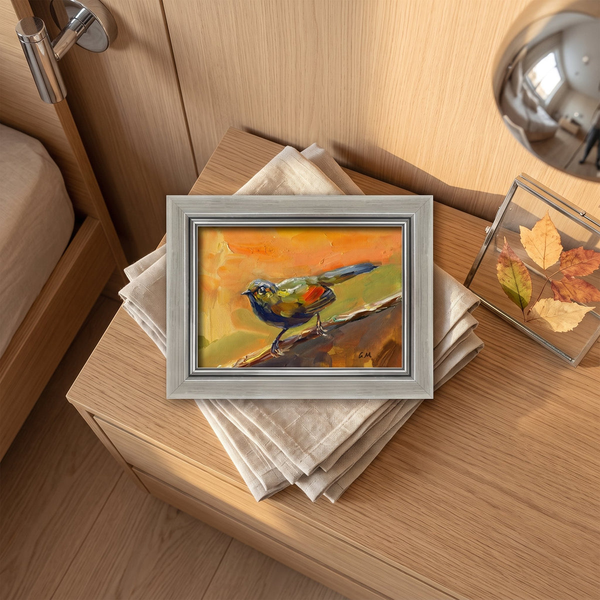 A vividly painted bird perched on a branch, rendered in bold orange, green, and blue hues, sits framed on a wooden nightstand beside folded linen and autumn leaves.