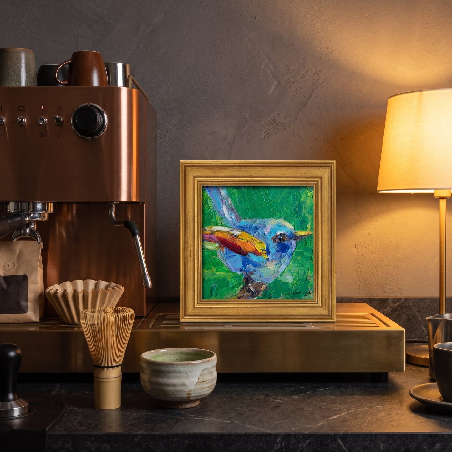 A colorful expressionist bird painting in a gold frame sits on a brass counter beside a coffee machine, glowing under warm lamplight.