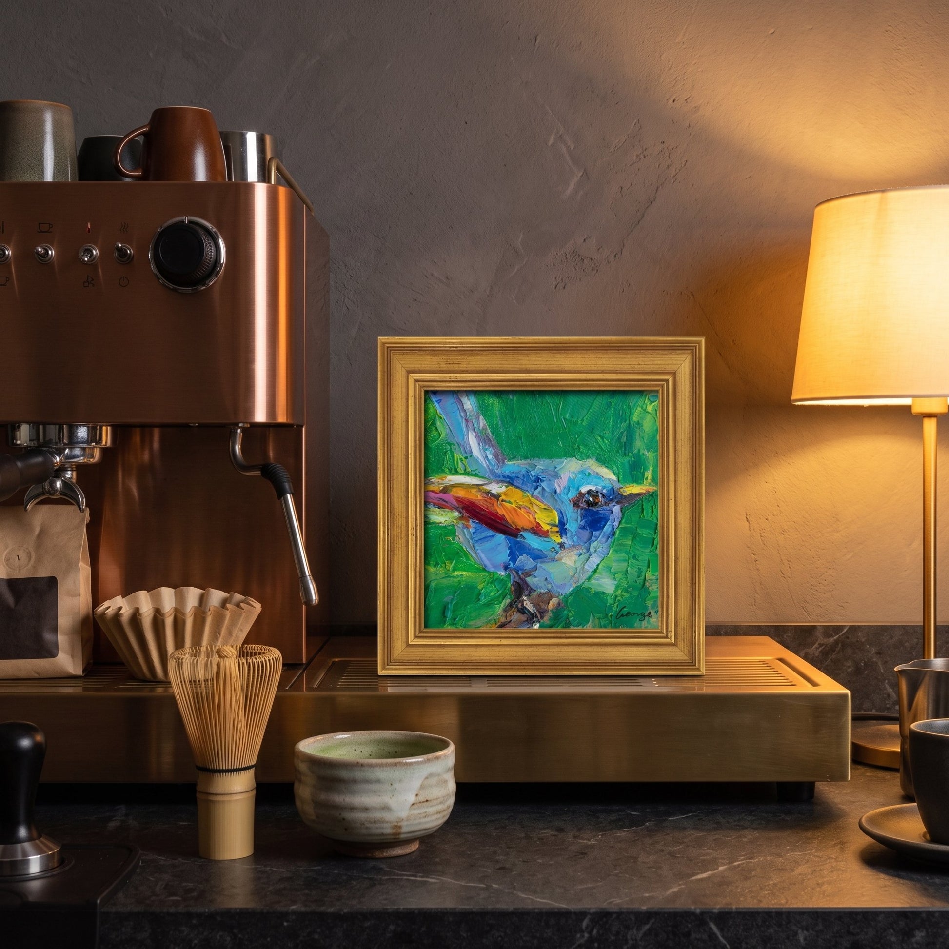 A colorful expressionist bird painting in a gold frame sits on a brass counter beside a coffee machine, glowing under warm lamplight.