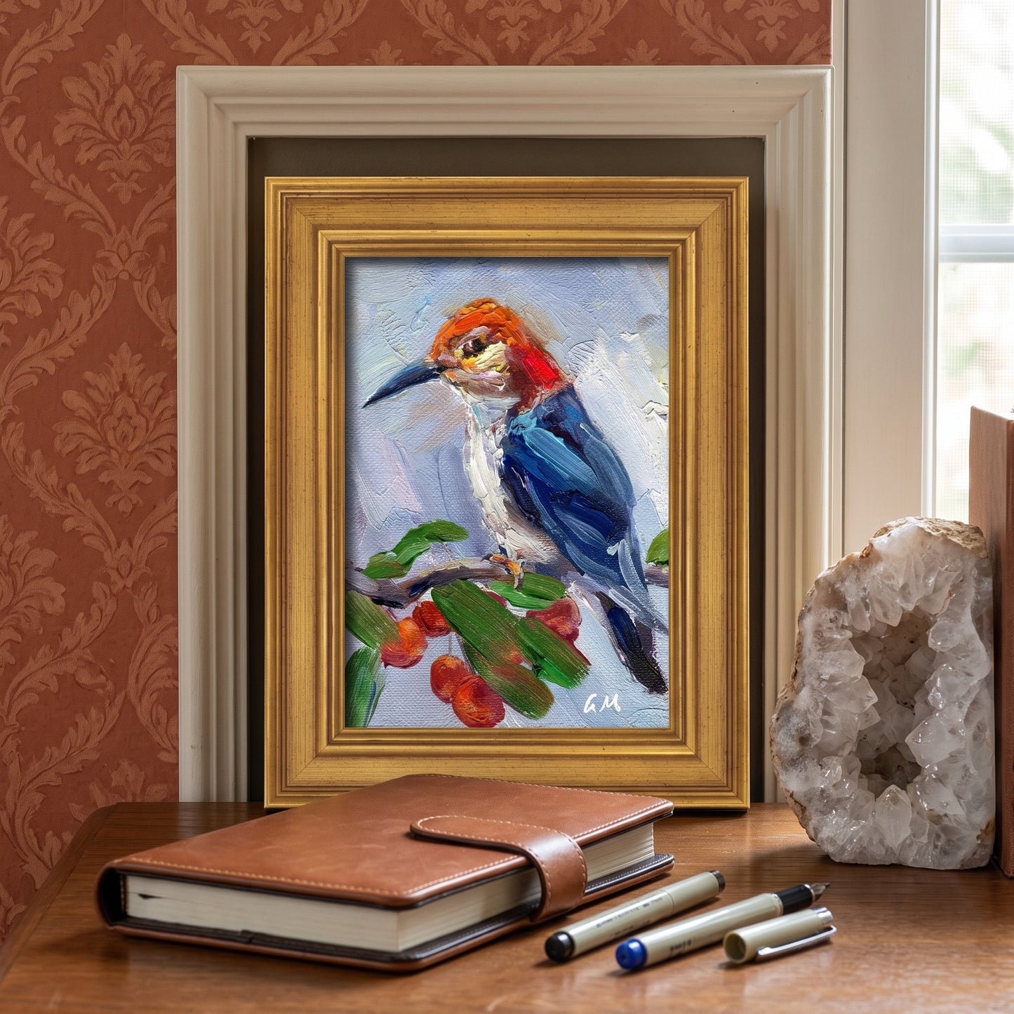 A vividly painted bird perched on berries in a gold-framed canvas sits on a wooden desk beside a leather journal and geode, bathed in soft daylight.