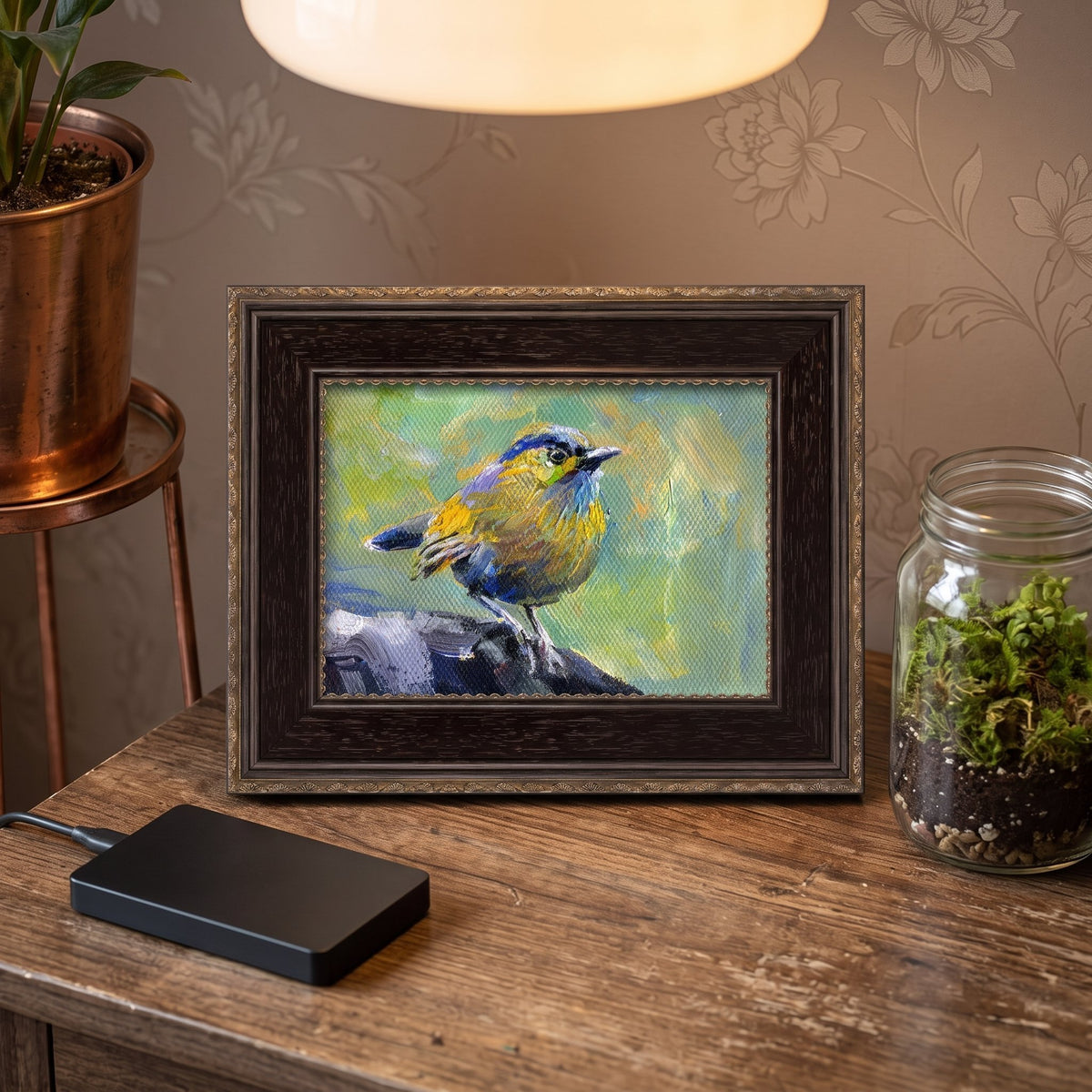 A vividly painted bird in blues and yellows rests on stone, displayed in a dark ornate frame atop a rustic wooden desk beside a terrarium and soft overhead lighting.