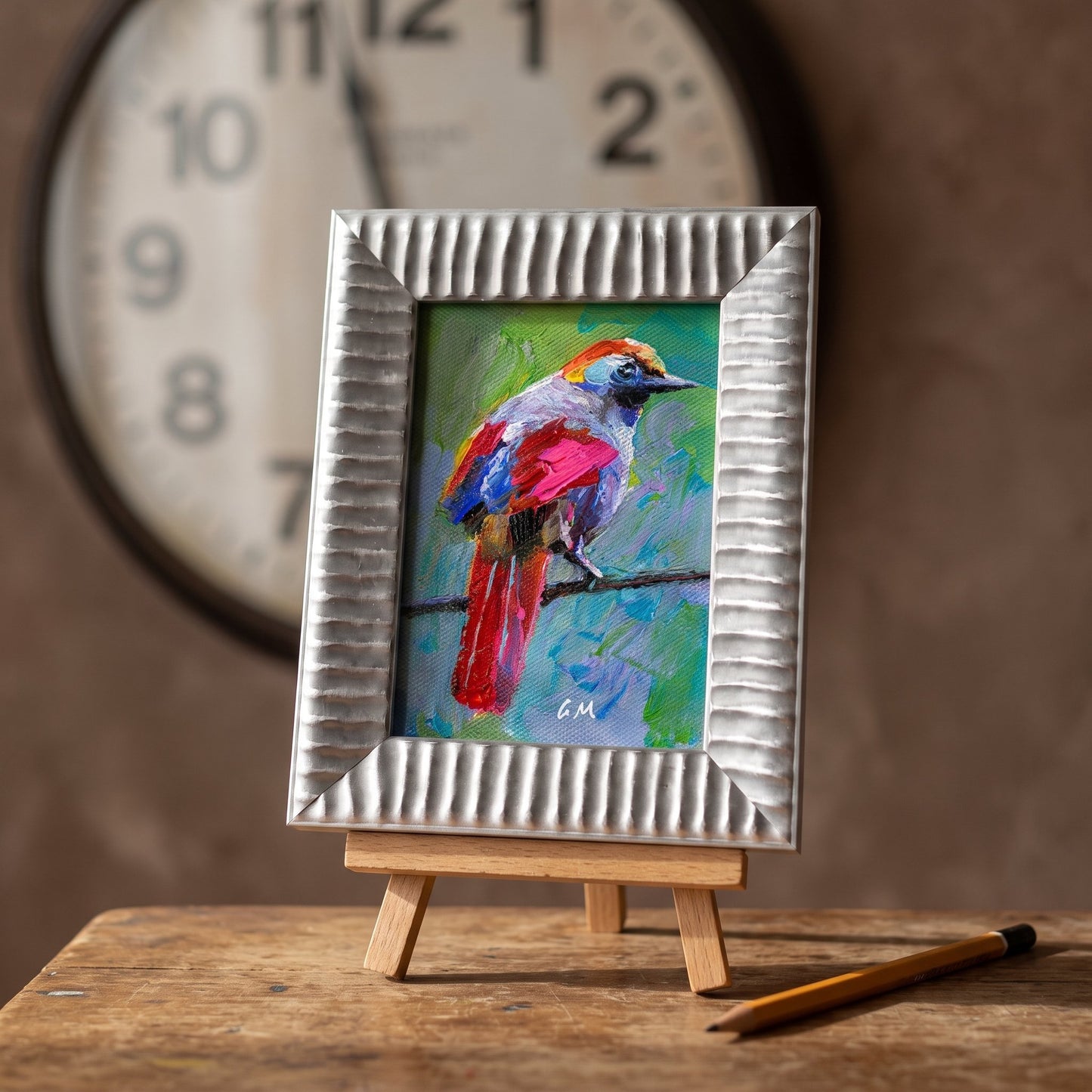 A vividly painted bird perched on a branch, rendered in bold reds, blues, and greens, rests on a wooden easel in a rustic study, framed in textured silver.