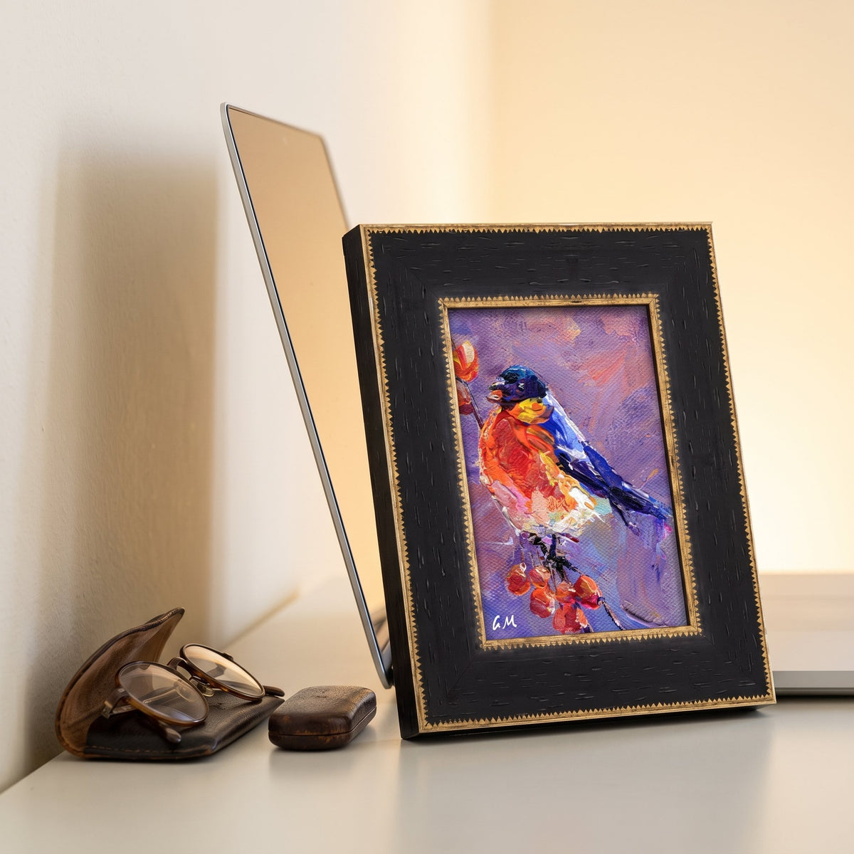 A vivid oil painting of a colorful bird perched on berries, initialed by the artist, rests in a black and gold frame beside a laptop and reading glasses on a sunlit desk.