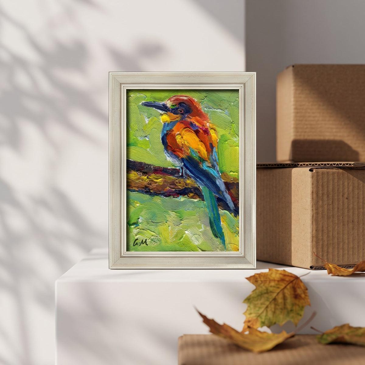 A vividly painted bird with fiery orange and deep blue feathers perches on a branch in an unframed canvas, displayed on a white pedestal beside autumn leaves and cardboard boxes.
